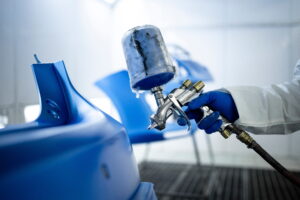 A person in protective gear spray-paints a blue car part in a workshop. The focus is on the paint gun and vibrant blue color, conveying precision.