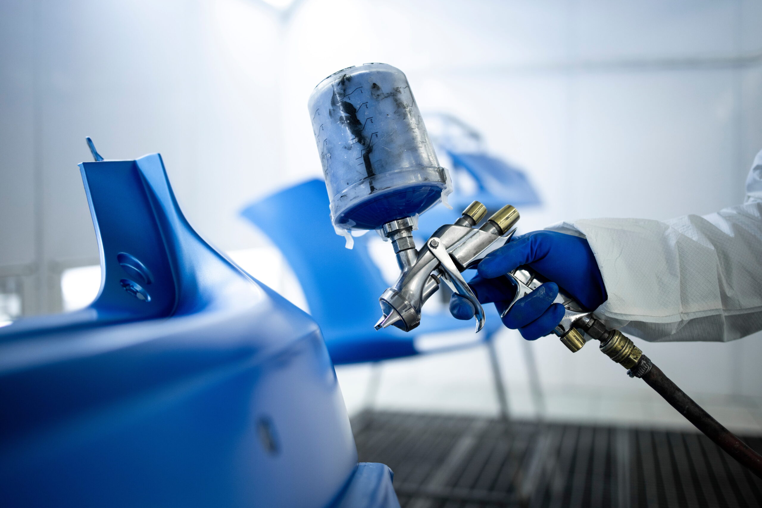 A person in protective gear spray-paints a blue car part in a workshop. The focus is on the paint gun and vibrant blue color, conveying precision.