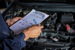 Mechanic in dark uniform holds a clipboard and pen while inspecting a car engine. The tone is focused and professional, emphasizing attention to detail.