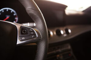 Close-up of a car's steering wheel with perforated leather. Showcases cruise control buttons, blurred dashboard, and gauge, conveying luxury and modern design.