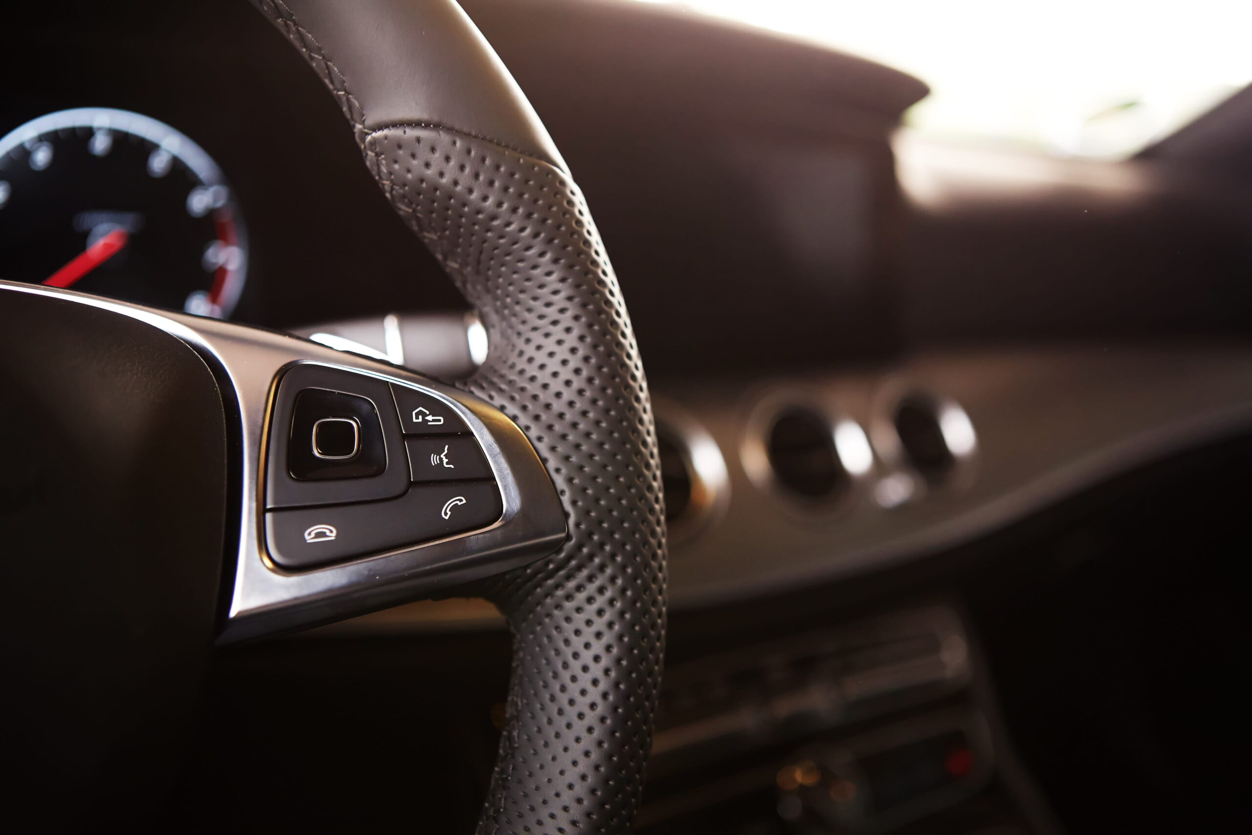 Close-up of a car's steering wheel with perforated leather. Showcases cruise control buttons, blurred dashboard, and gauge, conveying luxury and modern design.