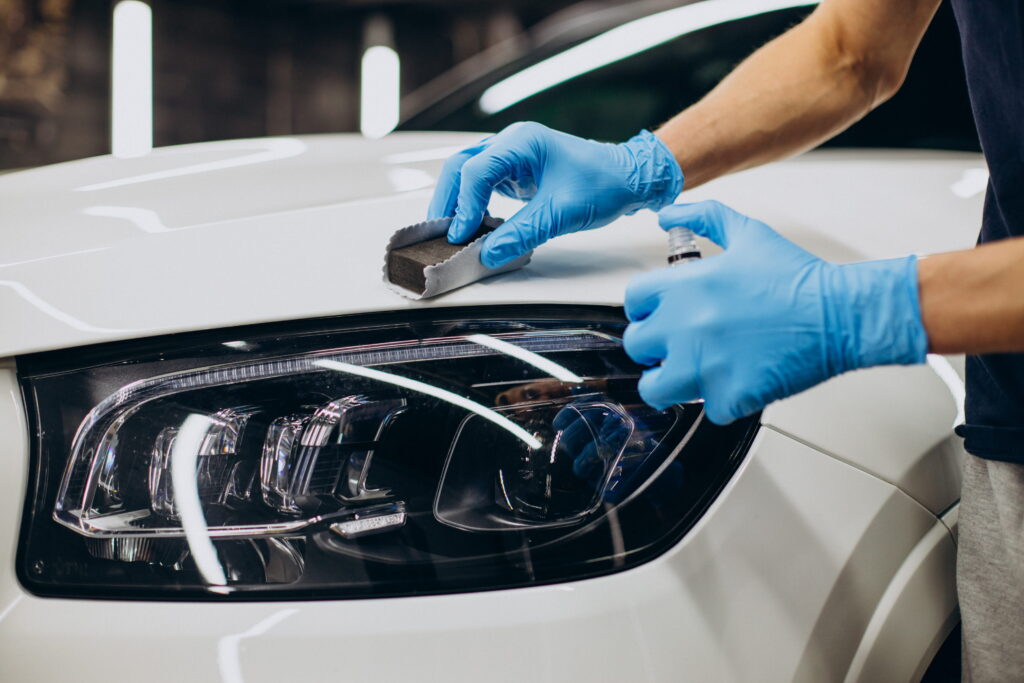 A person with blue gloves applies polish to a white car's headlight using a sponge, highlighting care and maintenance in an auto workshop setting.