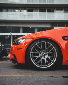 Close-up of a bright orange sports car side view, focusing on a sleek wheel with a BMW logo. The setting is urban, with a modern glass building in the background.
