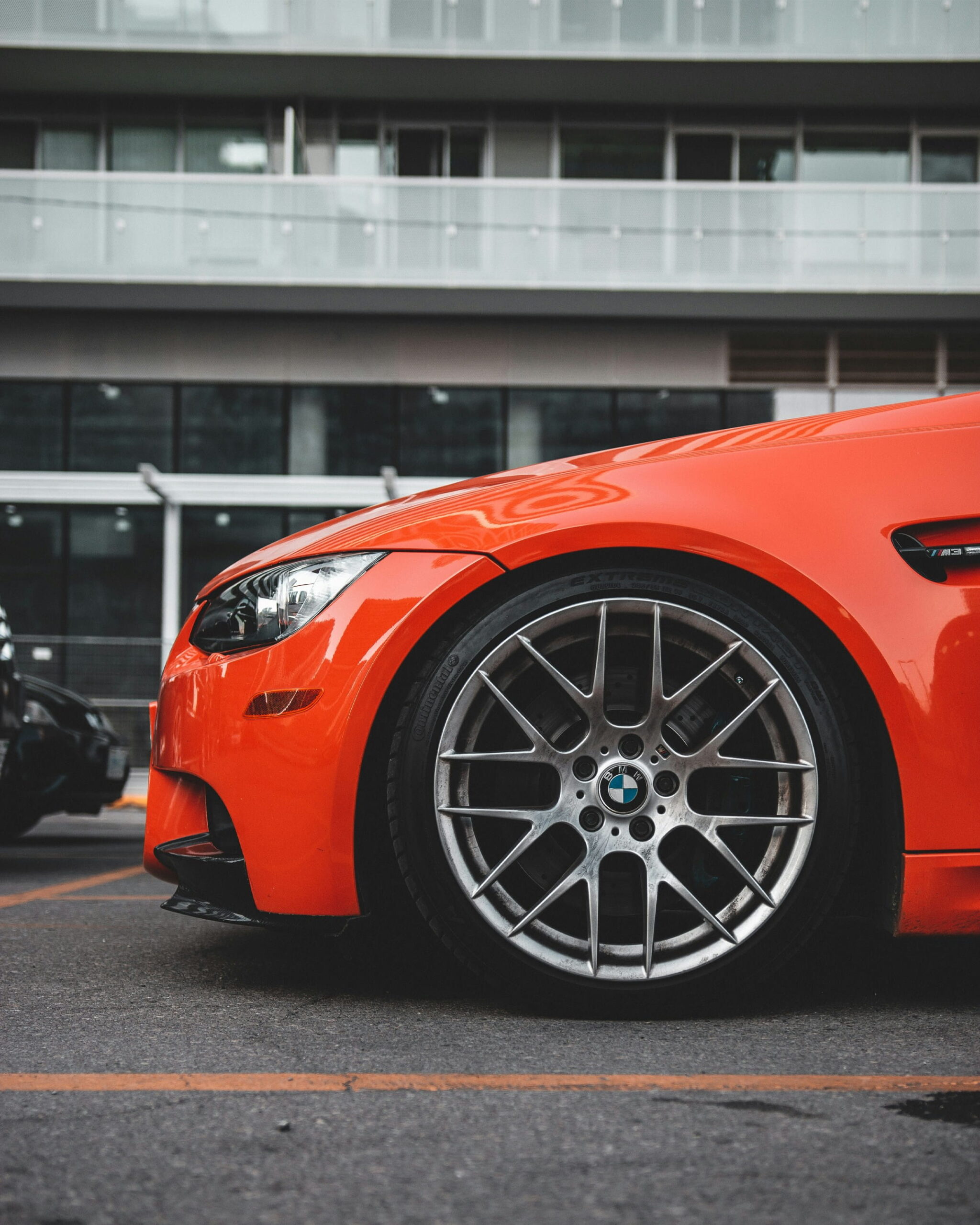 Close-up of a bright orange sports car side view, focusing on a sleek wheel with a BMW logo. The setting is urban, with a modern glass building in the background.