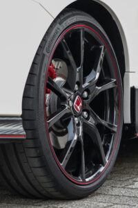 Close-up of a car's black alloy wheel with sleek red accents. The tire tread is visible, and the background is a section of a shiny white car body.