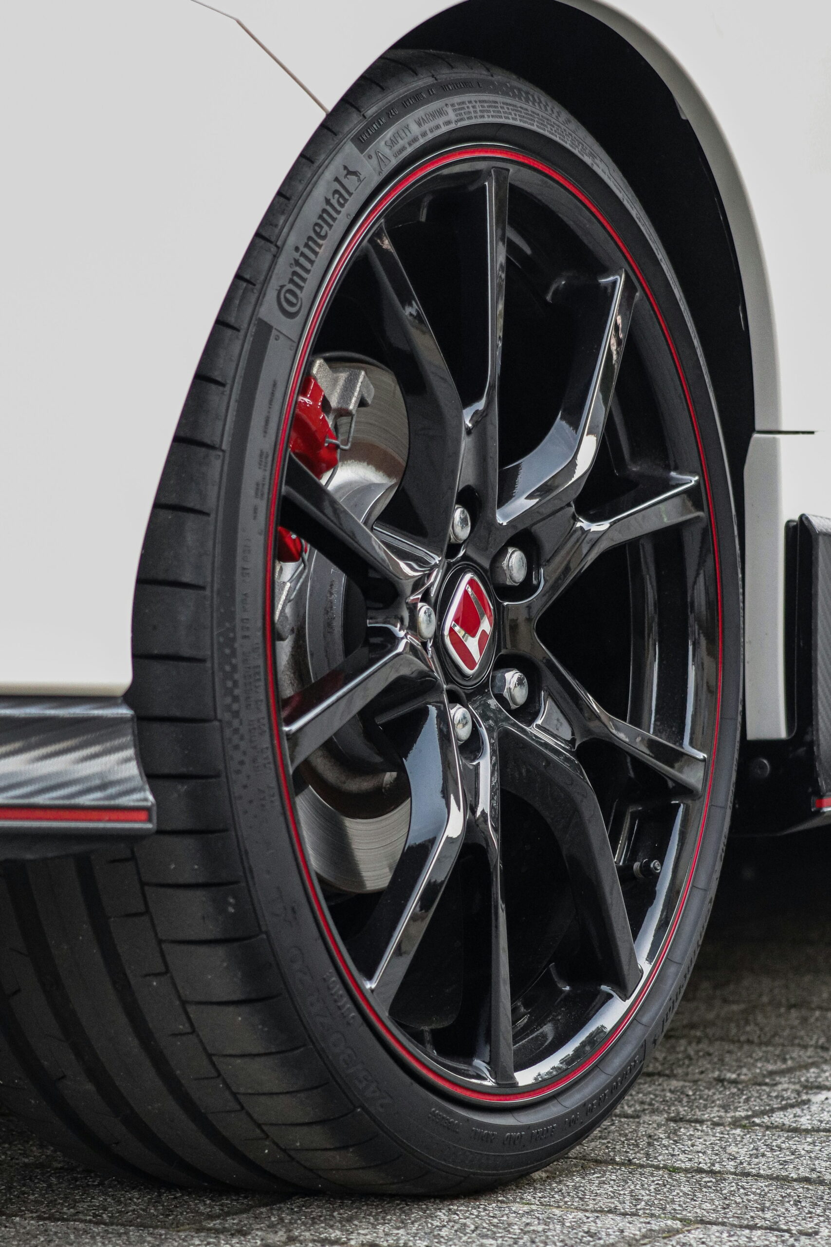 Close-up of a car's black alloy wheel with sleek red accents. The tire tread is visible, and the background is a section of a shiny white car body.