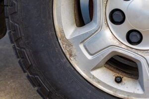 Close-up of a car wheel showing a dirty, weathered silver rim with visible rust and grime. The black rubber tire appears worn. The tone is neglected.