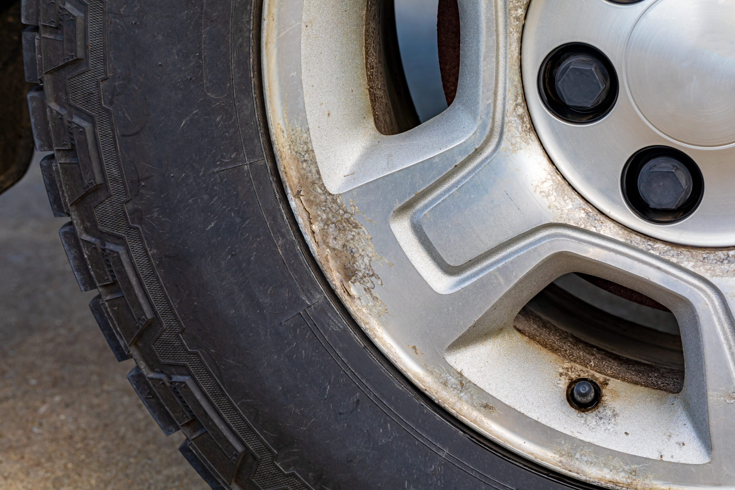 Close-up of a car wheel showing a dirty, weathered silver rim with visible rust and grime. The black rubber tire appears worn. The tone is neglected.